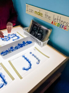 Preschool light table with a sign reading “I can make the letters” and a student using blue glass beads to trace the letter E