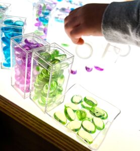 Glass beads in a container on a preschool light table with a child’s hand reaching to explore.