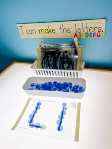 Preschool light table with a sign that says “I can make the letters” and a container of blue glass beads in front of a tracing sheet with the letter L