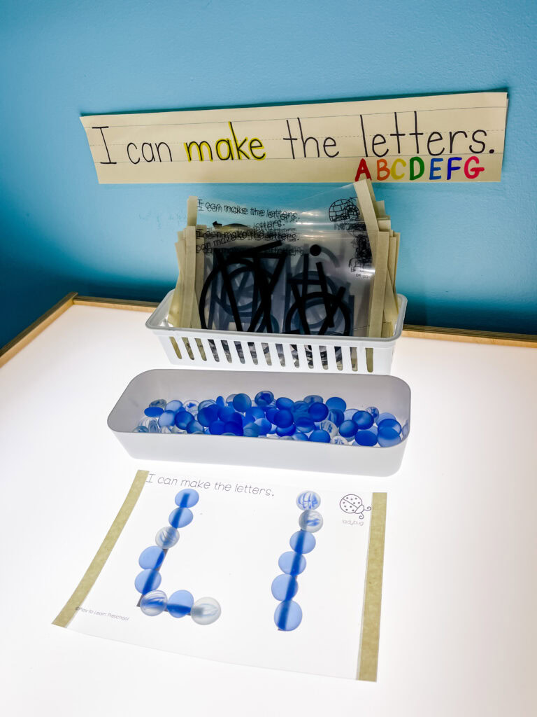 Preschool light table with a sign that says “I can make the letters” and a container of blue glass beads in front of a tracing sheet with the letter L