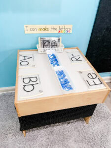 Preschool light table with a sign that says “I can make the letters,” four alphabet tracing sheets, and blue glass beads in the center