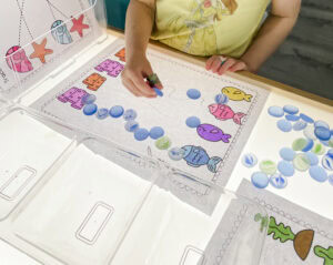Preschool student at a light table using glass beads to trace a pre-writing path from a fish to coral