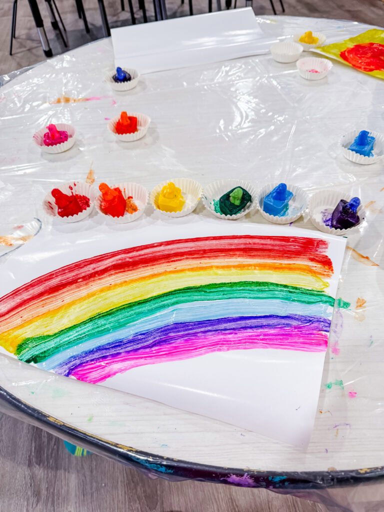 A preschool art table with frozen penguin paint cubes set in paper cups around a painted rainbow created with melting ice cube paints.