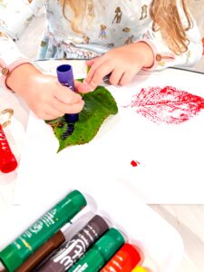 Preschool child painting a green leaf with a paint stick to create a nature print on paper.