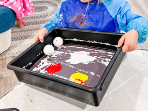 Preschool child using golf balls to roll paint across paper in a process art activity.