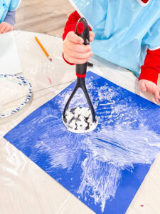 Preschool child stamping paint onto blue paper using a potato masher as an alternative painting tool.