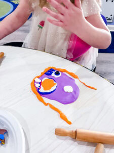 Purple Model Magic clay base formed into a face, decorated with orange clay hair and white and blue clay pieces for eyes and features.