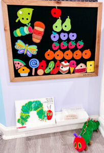 Black felt board with colorful pieces from The Very Hungry Caterpillar by Eric Carle, with the book and a caterpillar puppet displayed below.