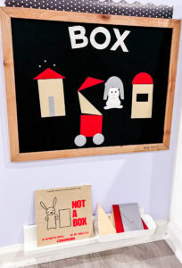 Black felt board with brown felt pieces and red accents; Not a Box book by Antoinette Portis sits on a shelf below.