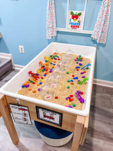 A sensory table filled with oats, plastic fruit pieces, colorful tweezers, and clear plastic jars arranged for sorting activities.