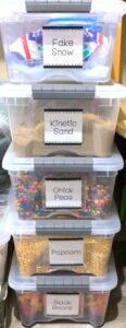 Stacked clear plastic tubs filled with sensory materials like fake snow, kinetic sand, chickpeas, popcorn, and black beans
