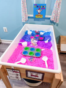 Pink, purple, and blue foam line a preschool sensory table. Measuring cups, cupcake tins, and birthday candles are on top.