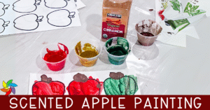 Scented apple painting setup with red, yellow, green, and brown paint in small cups, a container of cinnamon, and apple outline printables on a table.