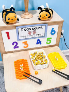 Counting bees math center setup with silicone honeycomb trays, plush bee toys, tongs, and a basket of bee counters on a preschool classroom table.