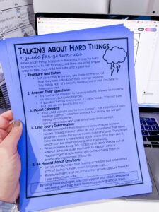 A hand holding a blue preschool parent handout titled “Talking About Hard Things: a guide for grown-ups.” The sheet provides supportive tips for talking to children about difficult or scary events, including sections on listening, answering questions, modeling calmness, limiting scary information, and being honest about emotions.
