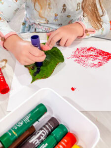 Preschooler using a purple tempera paint stick to color a green leaf while a red leaf print is visible on the paper.