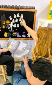 Teacher and preschool child using a felt board to retell Ten Timid Ghosts with ghost-shaped felt pieces while reading the book together.