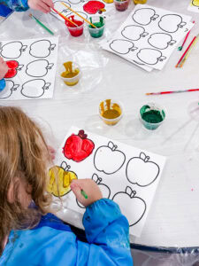 Preschool child painting apple outlines with yellow cinnamon-scented paint at an art table.