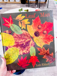 Close-up of the book Leaf Man by Lois Ehlert held in a preschool classroom with fall leaves and art materials in the background.