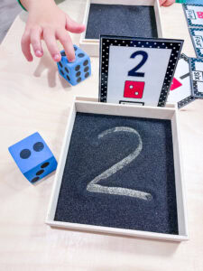 Roll and write math activity - Preschooler’s hand reaching for a foam die next to a black sand tray with the number 2 written in it and a matching number card nearby.