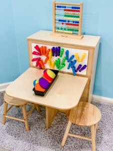 Preschool math center with an abacus on top, colorful chip clips arranged by color on a magnetic board, and a set of matching colored coasters on the table with two small stools.