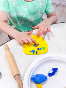 A preschool child pressing small colorful clay shapes onto a flattened yellow clay base as part of an art project.