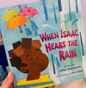 Close-up of the picture book When Isaac Hears the Rain by Julie Thompson, illustrated by Leah Giles. The cover shows a smiling child with stylized umbrellas and raindrops in the background.