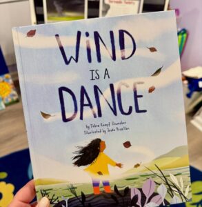 Cover of the book Wind Is a Dance by Debra Kempf Shumaker, showing a child standing in a breezy field with leaves swirling in the air.