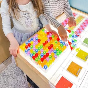 Two preschool children use colorful translucent pegs to create patterns and designs on a light-up pegboard placed on a light table.