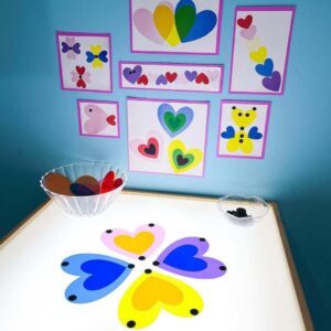 A light table is set up with colorful translucent heart shapes arranged in a flower pattern. A bowl holds more heart pieces. The wall behind displays examples of heart art made with similar shapes.