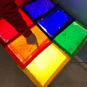 A child’s finger points to a colorful squish bag filled with yellow water beads on a light table. Other squish bags in red, orange, blue, and green are also glowing brightly on the table.