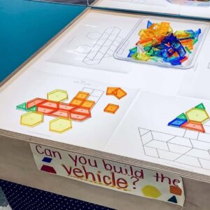 A preschool light table displays printable vehicle templates and a tray of colorful translucent pattern blocks. Some vehicles are partially built using the blocks.