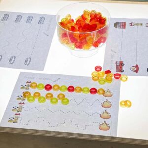A preschool light table displays tracing line worksheets with colorful translucent counters placed along the dotted lines. A bowl of orange, red, and yellow counters sits nearby.