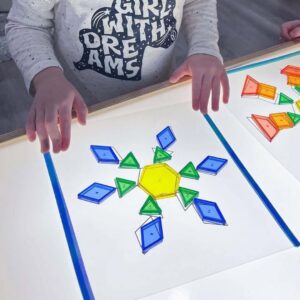 A child arranges translucent blue, green, and yellow pattern blocks on a light table to form a symmetrical snowflake shape.