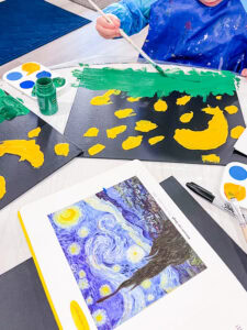 Child in a blue smock painting a Starry Night-inspired scene with green and yellow paint on black paper, with an art print of Van Gogh’s “Starry Night” nearby.