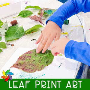 Preschooler pressing a painted leaf onto paper during a leaf print art activity, with green and red leaves and paint sticks visible on the table.