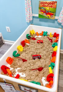 Alphabet sensory bin with kinetic sand, colorful plastic letters, red scoops, and a toy fire truck, set up with the book Alphabet Rescue.
