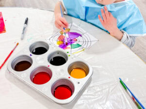 A preschool student works on a spider directed drawing, painting a black-lined spider web with bright liquid watercolors. A muffin tin holds colorful paint, and art supplies are scattered on the table.