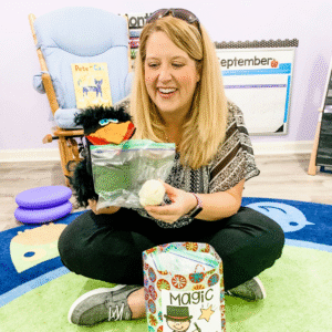 Teacher sitting on a classroom rug holding a puppet and a bag with a ball of dough during a preschool circle time activity.