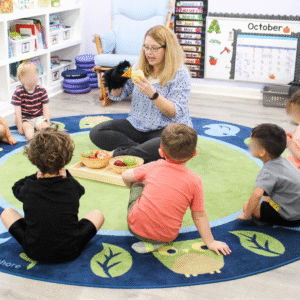 Teacher leading a preschool circle time with a puppet and a tray of apples as children sit on a classroom rug.