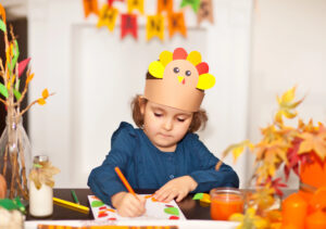 Preschool child wearing a turkey headband creating a Thanksgiving craft with crayons and colorful paper leaves.