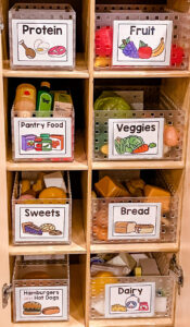 Preschool dramatic play kitchen cabinet organized with labeled bins for pretend food groups including protein, fruit, veggies, and dairy.
