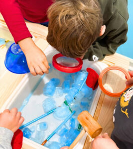 Ice cube sensory table with magnifying tools, paintbrushes, and water for preschool winter exploration.