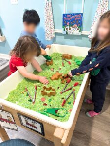Winter sensory table filled with green dyed rice, gingerbread shaped containers, tongs, scoops, and small holiday accents.