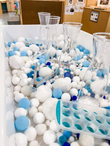 Winter themed sensory bin filled with blue and white pom poms, tongs, and clear containers for fine motor practice.