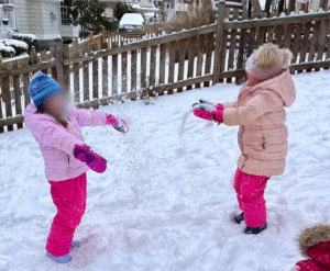 winter science observations: Two children in winter coats, hats, and mittens playing with snow outside, tossing snow into the air in a snowy backyard setting.