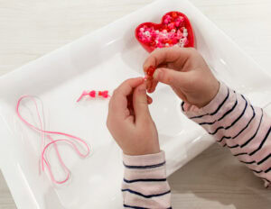 Child threading pink and red beads onto a string as part of a fine motor lacing activity.