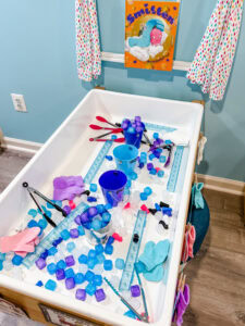 Winter sensory table with plastic ice cubes, buckets, tongs, rulers, and mittens arranged for scooping and transferring activities.