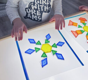 Children using translucent pattern blocks on a light table to build winter-themed shapes, including a large snowflake design made from colorful geometric pieces.