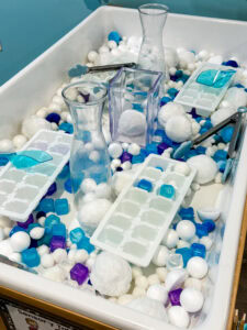 Winter-themed sensory bin filled with cotton snowballs, plastic ice cubes, scoops, tongs, pitchers, and ice cube trays for preschool fine motor play.